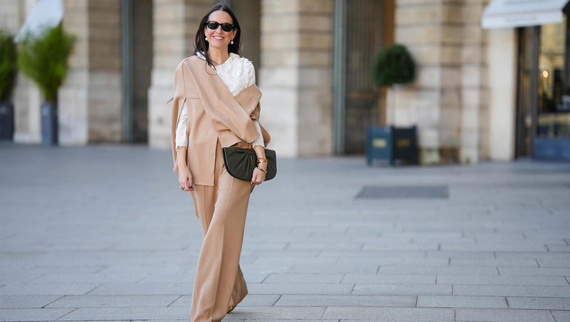 Alba Garavito Torre wears black sunglasses from Ray Ban, white large pearls earrings, a white shirt with cut-out lace pattern large collar, a beige felt / wool oversized cloak, matching beige wool / felt palazzo pants, a black shiny leather ruffled handbag, a gold Love bracelet from Cartier, gold bracelet, gold rings, brown leather pointed pumps heels shoes, during a street style fashion photo session, on October 28, 2021 in Paris, France.